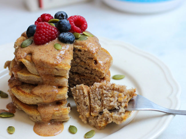 Stack of banana pancakes with berries, pumpkin seeds, and maple-almond sauce drizzled on top. 