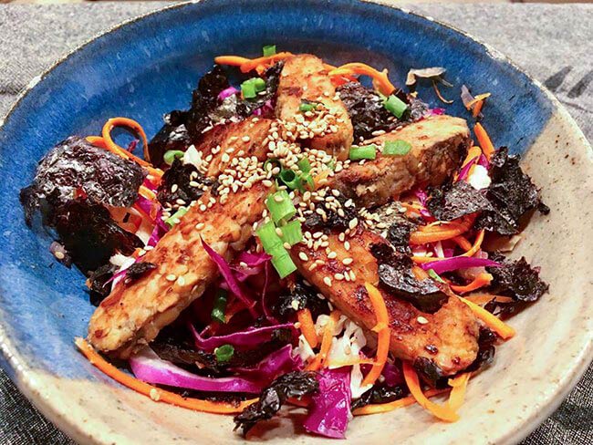 Barbecue tempeh salad garnished with seaweed, scallions, and sesame seeds in a clay bowl.