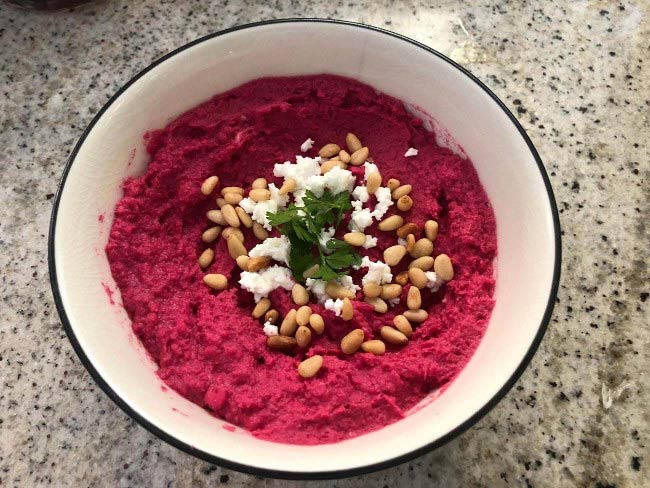 bowl of bright red hummus sprinkled with pine nuts and crumbled feta cheese and garnished with parsley