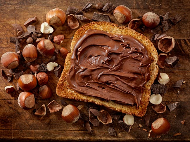Chocolate hazelnut spread on a piece of bread, surrounded by hazelnuts