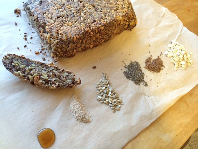A sliced loaf of whole-grain, gluten-free, bread on a fabric napkin, with small piles of grains and seeds.
