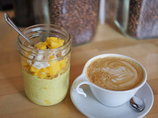 Golden milk overnight topped with mangos in a mason jar on a table next to a caffe latte in a white mug. 