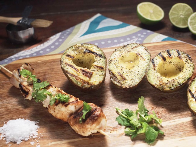 Skewer of chicken topped with cilantro leaves placed next to halved avocados striped with grill marks and sprinkled with pepper.