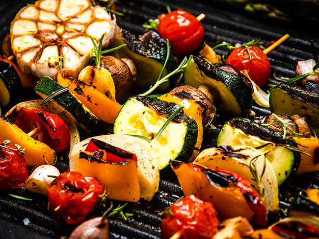 A flaming BBQ grill with skewers of fresh summer vegetables on top.