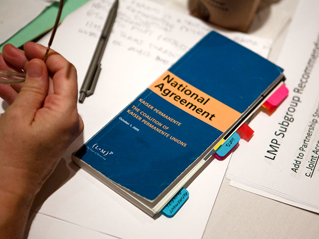 A man's hand is on a table holding a pair of glasses.  Next to the hand is a brochure titled: National Agreement Kaiser Permanente and The Coalition of Kaiser Permanente Unions.