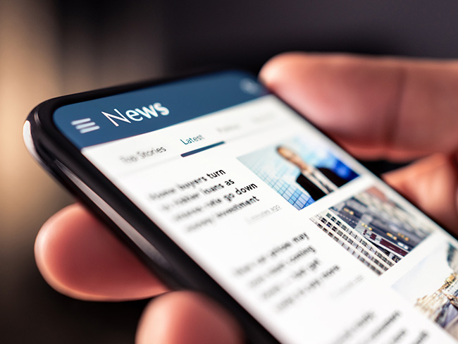 A man's hand holding a mobile phone, reading a news feed.