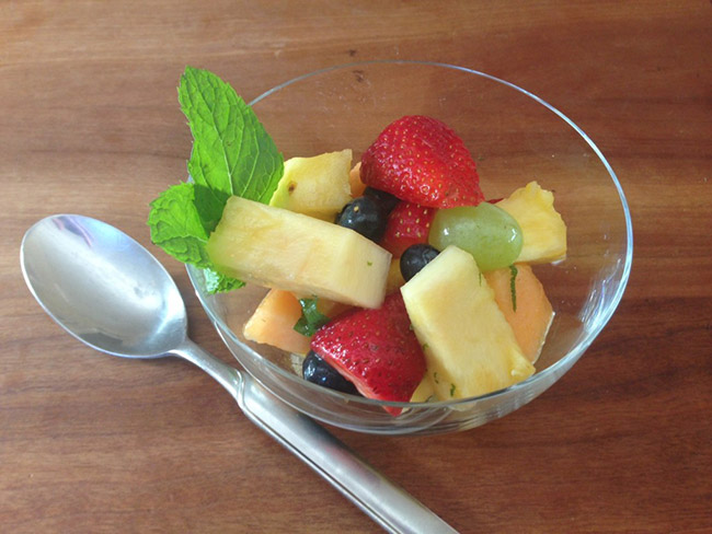 small glass bowl with grapes, blueberries, sliced strawberries, melon and pineapple chunks, and torn pieces of mint leaves