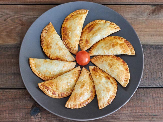 Overhead photo of 9 baked empanadas displayed in circle on gray plate, with cherry tomato in center