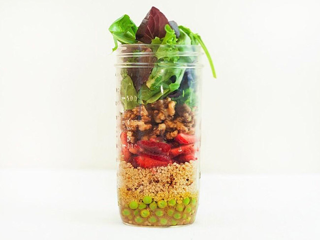 Layers of salad ingredients — lettuce, nuts, peas, quinoa — in a tall glass canning jar.