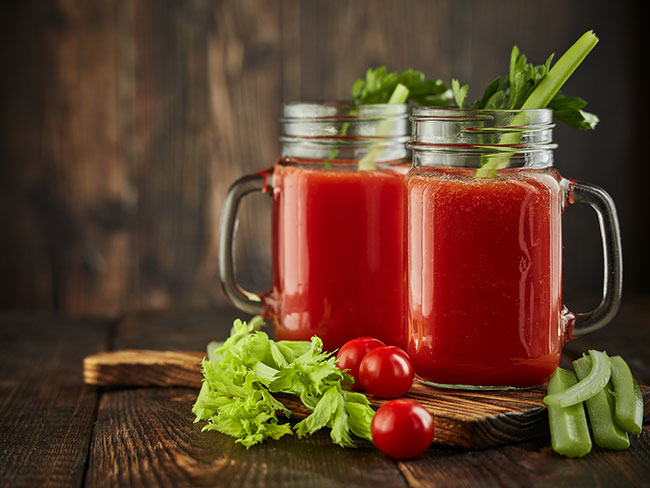 Two glasses of fresh vegetable juice, surrounded with celery, parsley and ripe tomatoes.