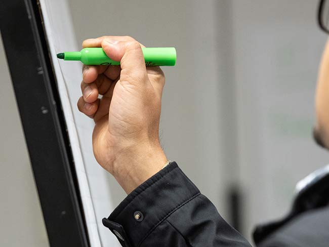  A person wearing a black jacket is writing on a whiteboard with a green marker.
