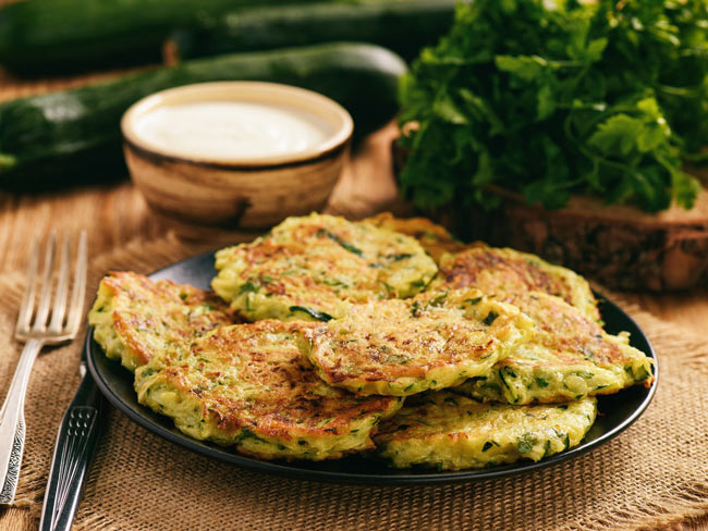 Zucchini fritters on wooden table, with greens, zucchini, and yogurt sauce in background.